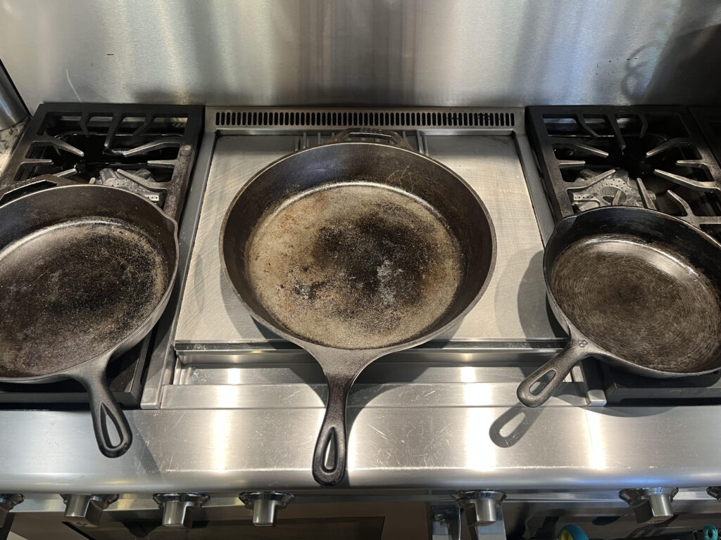 photo of three cast iron pans, small, medium, and large, sitting on a stainless steel stove or range