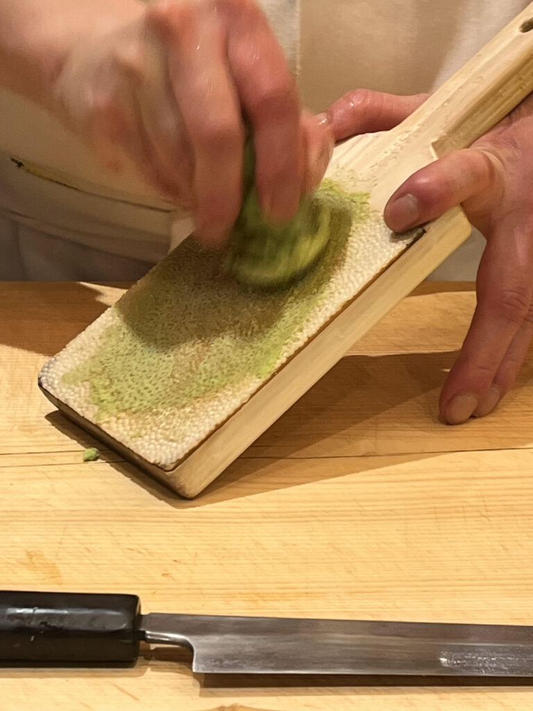 photo of a chef's hand grating fresh wasabi root on a shark skin grater made for grating fresh wasabi or ginger