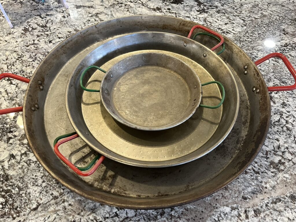 photo of three steel pans for cooking Spanish paella; the pans are three different sizes, are nested or stacked together, and have red or green handles
