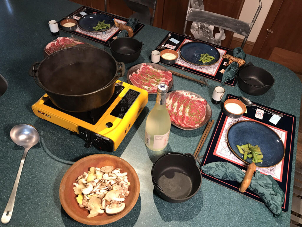 a typical dinner table setup for Japanese beef shabu shabu hotpot, with a Dutch oven iron pot on a butane stove burner with plates of sliced marbled beef for each guest, place settings and a bowl for each guest, and a bottle of sake; this photo illustrates the text