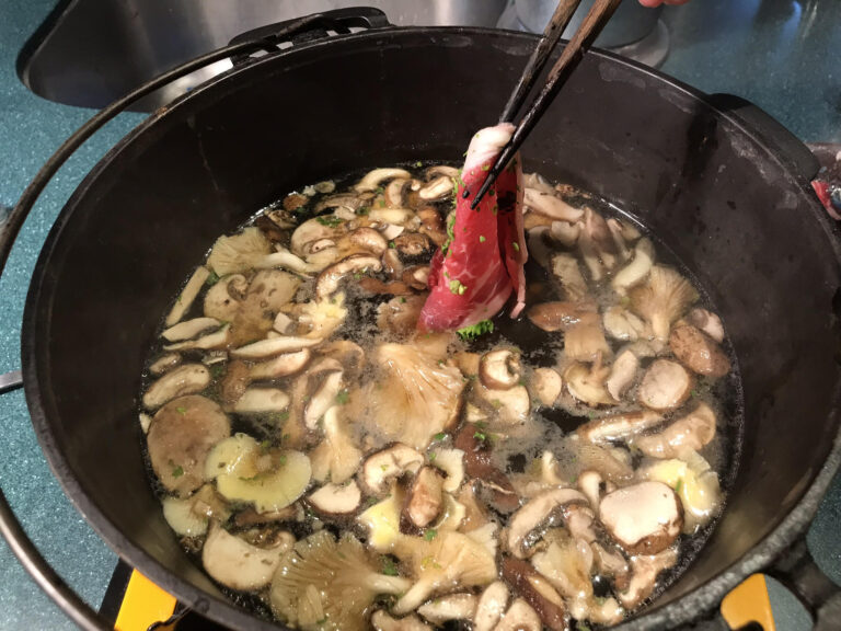 a cast iron pot with broth and mushrooms and chopsticks dipping a piece of beef in the broth