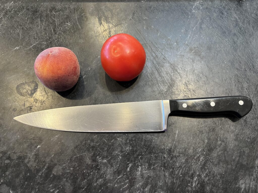 photo of a chef's knife on a marble board with a peach and a tomato