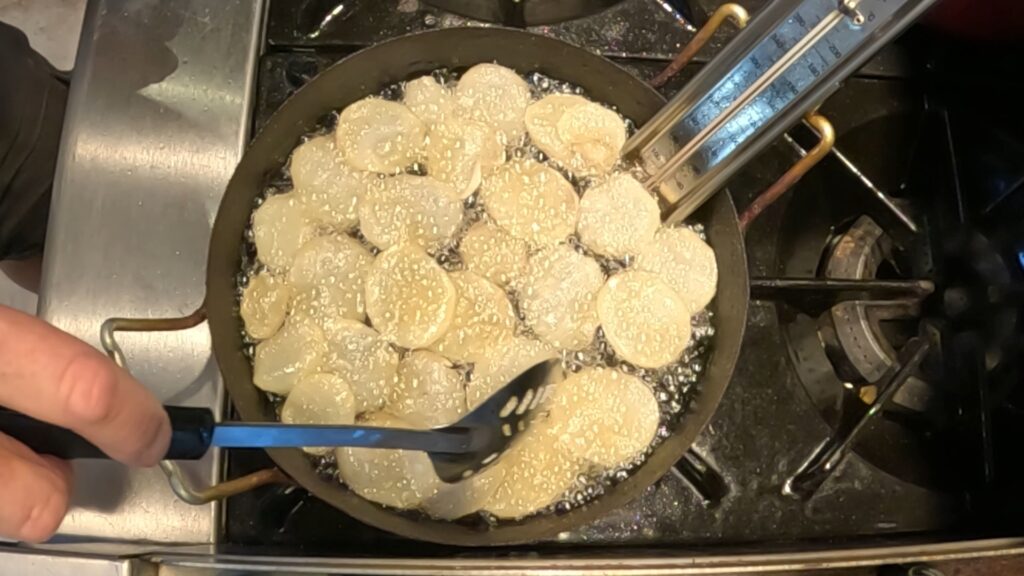 potato slices deep frying in a pot of oil with a slotted spoon and thermometer