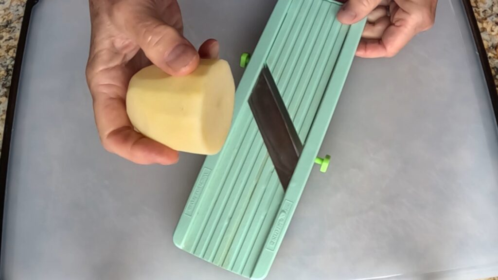 hands slicing a cut potato on a green mandoline