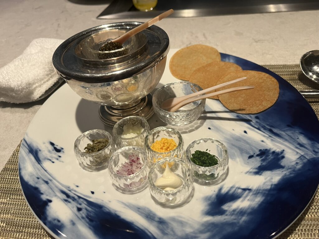 caviar service with a small bowl of caviar over ice, three blinis, and several small bowls with caviar accompaniments, chopped egg, chopped red onion, capers, etc.