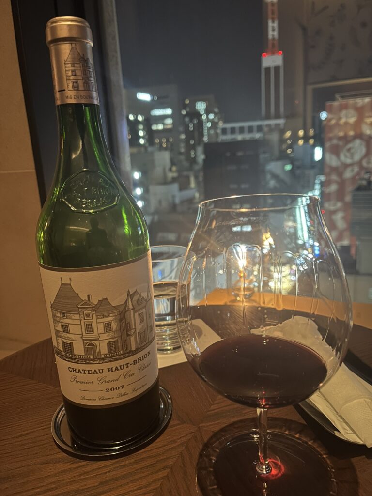 a bottle of Chateau Haut-Brion 2007 French red wine, a wine glass with some red wine in it, in front of a window with buildings in the backgroun