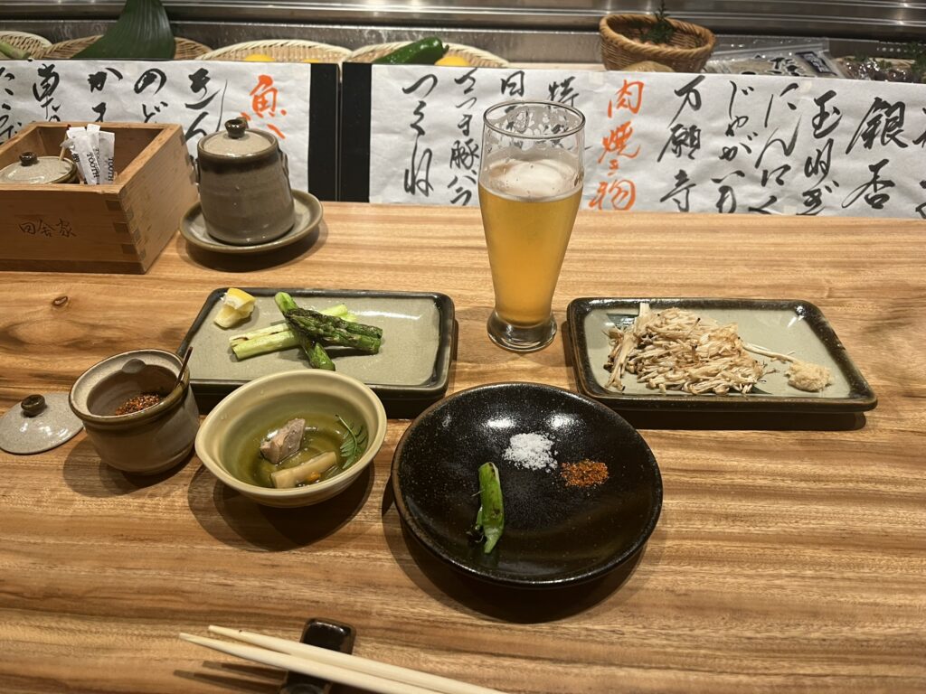 a plate of grilled asparagus, a plate of enoki mushrooms, a glass of beer, and several other dieshes