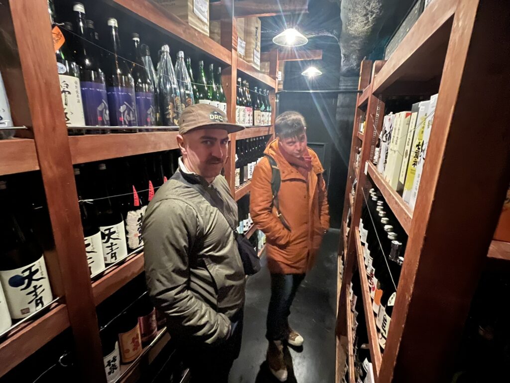 a sake shop with many bottles of sake on wooden shelves, with two customers